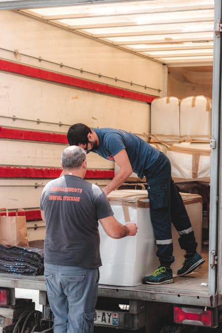 Two movers engaged in a home relocation process inside the back of a delivery truck. One worker, standing on the truck bed, is handling a large, wrapped, rectangular item secured with brown packing ta