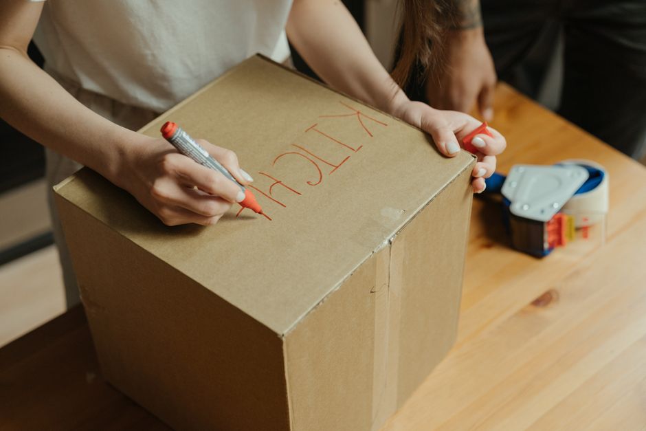 Close-up image of a cardboard moving box with a white paper label attached to the side. The label displays printed sections for 'Name,' 'Contents,' 'Order No.,' and 'Room,' with handwritten text showi