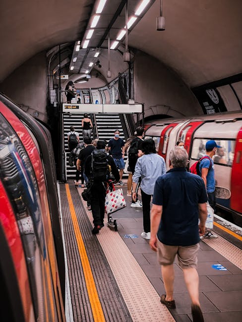 Image showing a busy underground station platform with people exiting a train on the right side. Several individuals are carrying backpacks, suitcases, or shopping bags, and some are wearing face mask